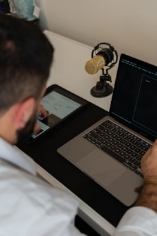 A person is sitting at a desk using a laptop and a tablet with a graphic and text visible on the screen. A gold-colored microphone is also set up nearby on the desk. The person is casually dressed and seems to be focused on their work.