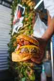 A close-up of a hand holding a large, juicy burger with layers of melted cheese, crispy bacon, and a thick beef patty sandwiched between toasted buns. The background contains blurred greenery and a partially visible sign, suggesting an outdoor setting or food truck environment.