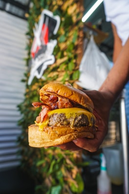A close-up of a hand holding a large, juicy burger with layers of melted cheese, crispy bacon, and a thick beef patty sandwiched between toasted buns. The background contains blurred greenery and a partially visible sign, suggesting an outdoor setting or food truck environment.
