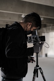 A person wearing a black jacket and gloves operates a professional camera mounted on a tripod. The surroundings suggest an industrial or urban setting with concrete and muted lighting.