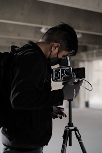 A person wearing a black jacket and gloves operates a professional camera mounted on a tripod. The surroundings suggest an industrial or urban setting with concrete and muted lighting.