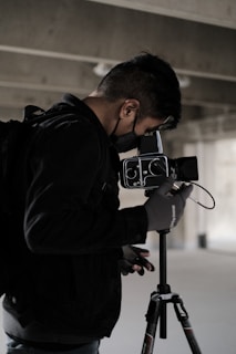 A person wearing a black jacket and gloves operates a professional camera mounted on a tripod. The surroundings suggest an industrial or urban setting with concrete and muted lighting.