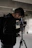 A person wearing a black jacket and gloves operates a professional camera mounted on a tripod. The surroundings suggest an industrial or urban setting with concrete and muted lighting.