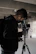 A person wearing a black jacket and gloves operates a professional camera mounted on a tripod. The surroundings suggest an industrial or urban setting with concrete and muted lighting.