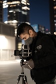 A photographer in a dark jacket and mask is adjusting a camera mounted on a tripod. The setting appears to be an urban night scene, with tall buildings in the background illuminated by artificial lights, creating a bokeh effect.