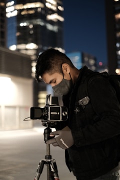 A photographer in a dark jacket and mask is adjusting a camera mounted on a tripod. The setting appears to be an urban night scene, with tall buildings in the background illuminated by artificial lights, creating a bokeh effect.