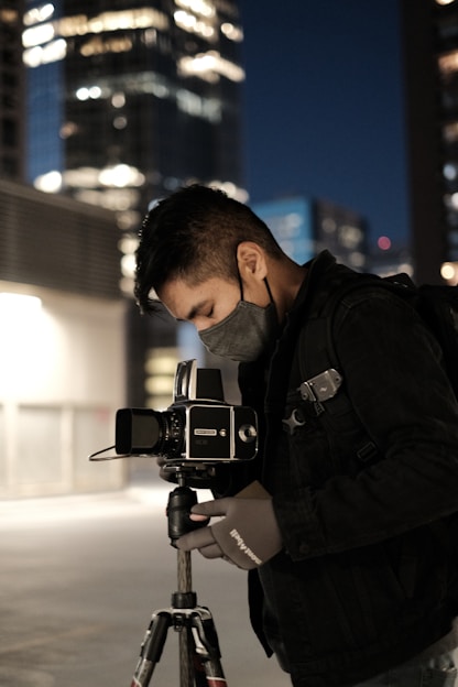 A photographer in a dark jacket and mask is adjusting a camera mounted on a tripod. The setting appears to be an urban night scene, with tall buildings in the background illuminated by artificial lights, creating a bokeh effect.