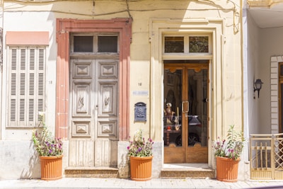 A quaint street view featuring two doors side by side, one wooden with detailed carvings and another with glass panes reflecting a car and a mannequin head wearing a hat. The facade is accented with a soft beige and terracotta frame. Potted plants with colorful flowers decorate the front, and a vintage-style wall-mounted lamp is visible.