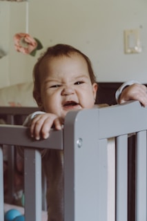 A baby stands in a crib, holding onto the railing with one hand. The child has a scrunched, playful expression on their face. Behind the baby, a soft toy mobile with a pink flower is just visible.