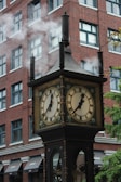 A vintage steam-powered clock with multiple faces displays time with Roman numerals. It emits visible steam through its chimneys. This clock is set against the backdrop of a red brick multi-story building with numerous windows. There are also some visible awnings on the building.
