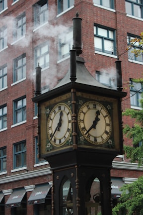 A vintage steam-powered clock with multiple faces displays time with Roman numerals. It emits visible steam through its chimneys. This clock is set against the backdrop of a red brick multi-story building with numerous windows. There are also some visible awnings on the building.