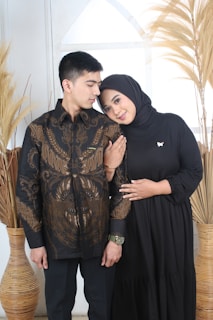 A couple is standing close together, with the woman resting her head on the man's shoulder. The man is wearing a black shirt with intricate brown patterns, while the woman is in a black dress and a black hijab. They are standing in front of a white background with dried plants in decorative vases on either side.