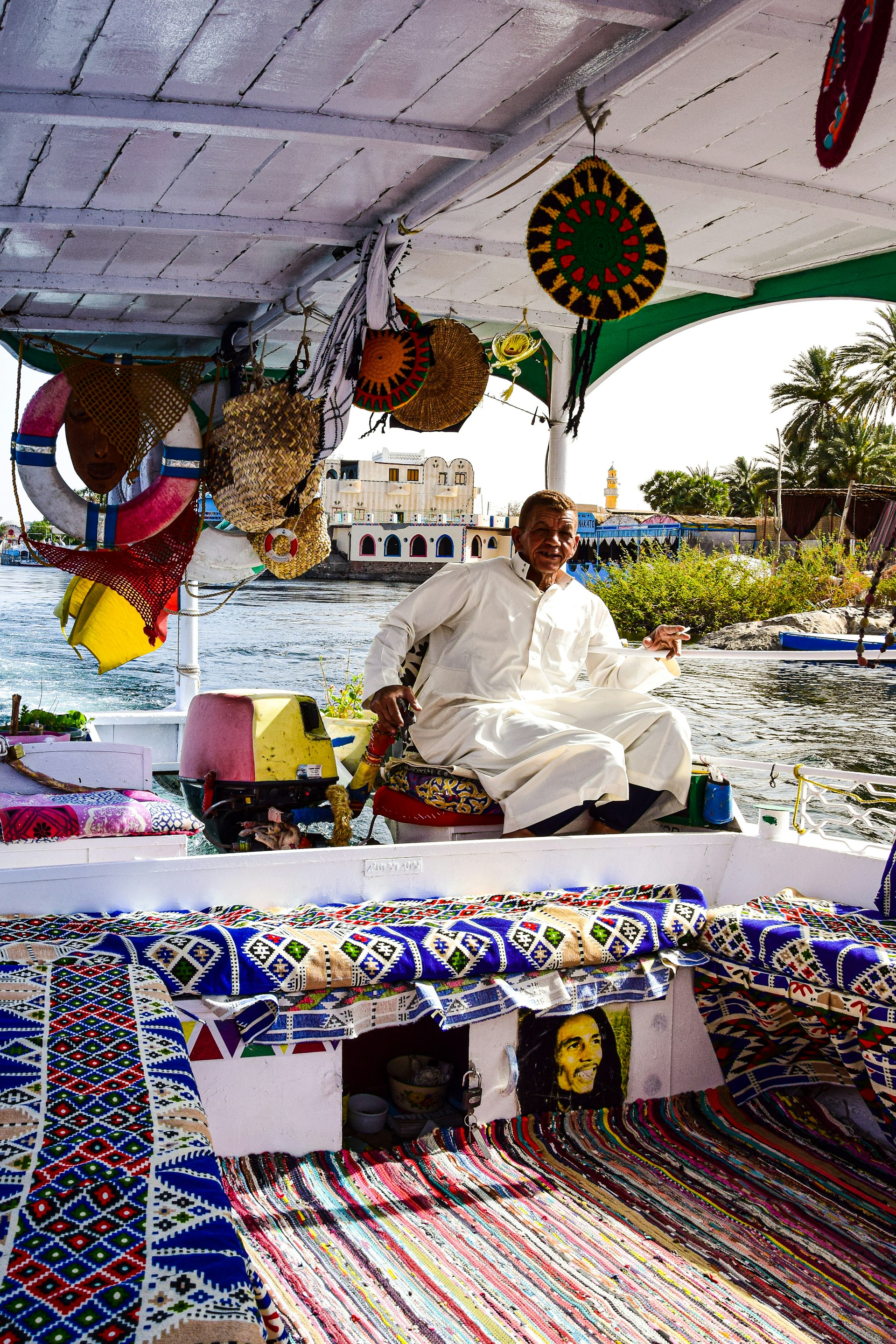 A man wearing a traditional white robe is sitting on a vibrant and colorful boat adorned with intricate patterned textiles. Various woven baskets and decorative items are hung around the boat's interior. The river flows calmly in the background, with buildings and lush greenery visible along the riverside.