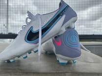 A pair of soccer cleats, primarily white with accents of blue, purple, and black. The cleats feature branding elements in contrasting colors, including a prominent logo and decorative designs. They are positioned on a concrete surface with a metal grid backdrop and a slightly overcast sky.