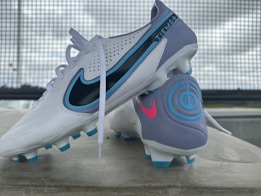 A pair of soccer cleats, primarily white with accents of blue, purple, and black. The cleats feature branding elements in contrasting colors, including a prominent logo and decorative designs. They are positioned on a concrete surface with a metal grid backdrop and a slightly overcast sky.