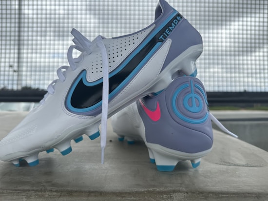 A pair of soccer cleats, primarily white with accents of blue, purple, and black. The cleats feature branding elements in contrasting colors, including a prominent logo and decorative designs. They are positioned on a concrete surface with a metal grid backdrop and a slightly overcast sky.