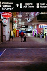 A person wearing a red jacket is walking down an airport terminal corridor with a rolling suitcase. Overhead signs indicate directions for wheelchairs, first class, TSA Precheck, ticketing, and gates. Various advertisements and a Coca-Cola vending machine are visible along the sides.
