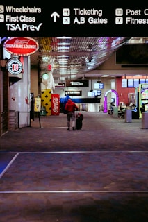 A person wearing a red jacket is walking down an airport terminal corridor with a rolling suitcase. Overhead signs indicate directions for wheelchairs, first class, TSA Precheck, ticketing, and gates. Various advertisements and a Coca-Cola vending machine are visible along the sides.