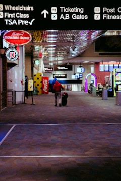 A person wearing a red jacket is walking down an airport terminal corridor with a rolling suitcase. Overhead signs indicate directions for wheelchairs, first class, TSA Precheck, ticketing, and gates. Various advertisements and a Coca-Cola vending machine are visible along the sides.