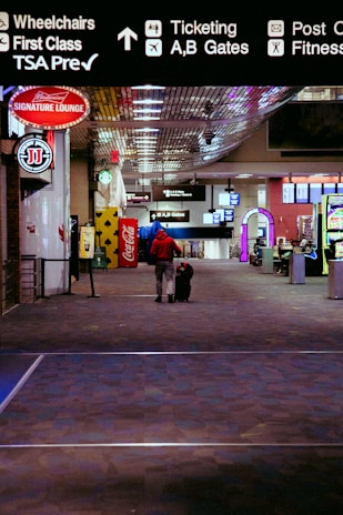 A person wearing a red jacket is walking down an airport terminal corridor with a rolling suitcase. Overhead signs indicate directions for wheelchairs, first class, TSA Precheck, ticketing, and gates. Various advertisements and a Coca-Cola vending machine are visible along the sides.
