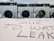 A row of industrial washing machines is visible in the background, while a cardboard sign in the foreground reads 'CLOSED DUE TO WAT LEAK' in handwritten letters.