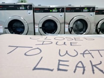 A row of industrial washing machines is visible in the background, while a cardboard sign in the foreground reads 'CLOSED DUE TO WAT LEAK' in handwritten letters.