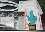 A window displays two USPS Priority Mail labels. One label features a colorful drawing of a cactus in a pot, while the other appears worn and partially covered by white scribbles on the glass.