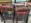 Several stacked wooden chairs are covered in splatters of paint in various colors, including red, black, blue, and yellow. Behind the chairs, there are assorted art objects and ceramics on a shelf, including figurines and colorful pottery. The setting appears to be an art studio or workshop.