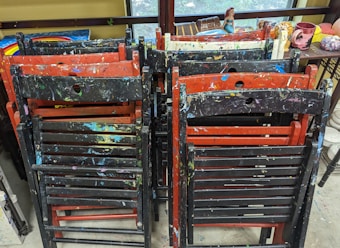 Several stacked wooden chairs are covered in splatters of paint in various colors, including red, black, blue, and yellow. Behind the chairs, there are assorted art objects and ceramics on a shelf, including figurines and colorful pottery. The setting appears to be an art studio or workshop.