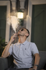 A person wearing sunglasses and a light-colored polo shirt lounges on a chair, exhaling a plume of smoke into the air. The background shows a dimly lit room with a glowing lamp on the wall.