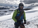 A person wearing a bright green and blue jacket is standing on a snow-covered mountain with climbing gear and ropes. The background shows rugged, snow-capped peaks under a cloudy sky.
