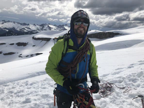 A rugged man wearing a waterproof jacket and insulated gloves, standing on a snowy mountain peak at sunrise.