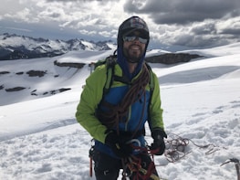 A person wearing a bright green and blue jacket is standing on a snow-covered mountain with climbing gear and ropes. The background shows rugged, snow-capped peaks under a cloudy sky.