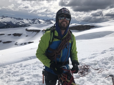 A person wearing a bright green and blue jacket is standing on a snow-covered mountain with climbing gear and ropes. The background shows rugged, snow-capped peaks under a cloudy sky.