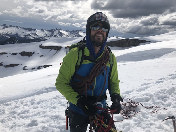 A person wearing a bright green and blue jacket is standing on a snow-covered mountain with climbing gear and ropes. The background shows rugged, snow-capped peaks under a cloudy sky.