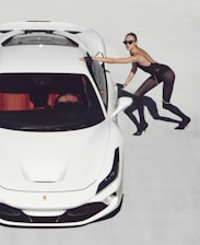 A woman in a stylish, fitted outfit leans against a sleek white sports car on a light background. She wears sunglasses and high heels, projecting a fashionable and confident demeanor.