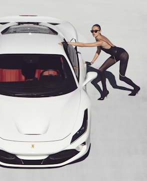 A woman in a stylish, fitted outfit leans against a sleek white sports car on a light background. She wears sunglasses and high heels, projecting a fashionable and confident demeanor.