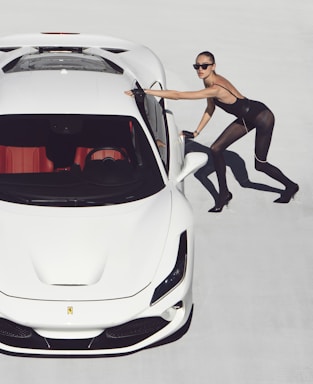 A woman in a stylish, fitted outfit leans against a sleek white sports car on a light background. She wears sunglasses and high heels, projecting a fashionable and confident demeanor.