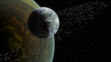 A large planet with a textured surface is positioned near the edge of the frame, with an abundance of smaller celestial bodies orbiting around it against a backdrop of space. The planet displays shades of green, brown, and yellow, creating a complex pattern. The surrounding space is dark, dotted with numerous scattered asteroids or moons.