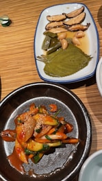 Two dishes are presented on a wooden table. The first dish on a white plate features sliced mushrooms, green peppers, whole garlic cloves, and a large leaf, all immersed in a light brown sauce. The second dish is in a dark bowl, showcasing a colorful salad with sliced cucumbers, onions, and carrots coated with a spicy red sauce and garnished with sesame seeds.