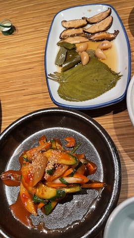 Two dishes are presented on a wooden table. The first dish on a white plate features sliced mushrooms, green peppers, whole garlic cloves, and a large leaf, all immersed in a light brown sauce. The second dish is in a dark bowl, showcasing a colorful salad with sliced cucumbers, onions, and carrots coated with a spicy red sauce and garnished with sesame seeds.