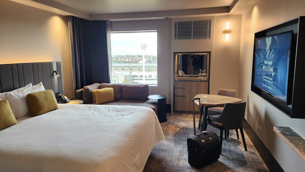 A modern hotel room features a large bed with white linens and mustard-colored pillows. There's a cozy sofa with similar cushion accents by the window, which provides a view of some outside structures. A round table with two chairs sits near a wall-mounted flat-screen TV displaying a welcome message. A piece of luggage is placed on the floor next to the table. The room has soft lighting and carpeted flooring, creating a comfortable and inviting atmosphere.