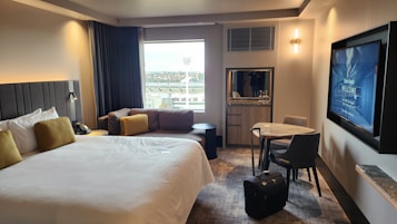 A modern hotel room features a large bed with white linens and mustard-colored pillows. There's a cozy sofa with similar cushion accents by the window, which provides a view of some outside structures. A round table with two chairs sits near a wall-mounted flat-screen TV displaying a welcome message. A piece of luggage is placed on the floor next to the table. The room has soft lighting and carpeted flooring, creating a comfortable and inviting atmosphere.