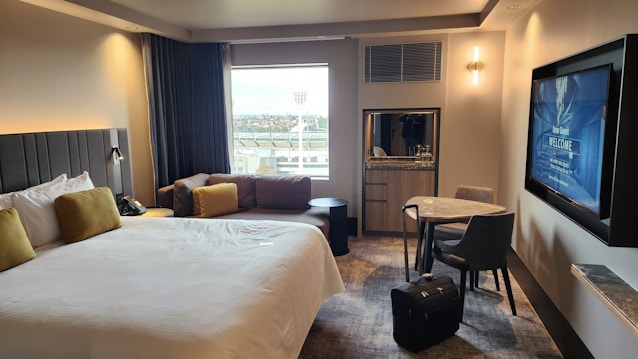 A modern hotel room features a large bed with white linens and mustard-colored pillows. There's a cozy sofa with similar cushion accents by the window, which provides a view of some outside structures. A round table with two chairs sits near a wall-mounted flat-screen TV displaying a welcome message. A piece of luggage is placed on the floor next to the table. The room has soft lighting and carpeted flooring, creating a comfortable and inviting atmosphere.