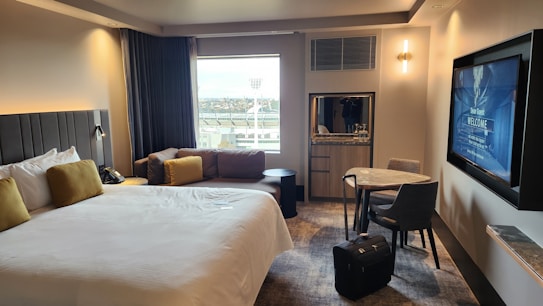 A modern hotel room features a large bed with white linens and mustard-colored pillows. There's a cozy sofa with similar cushion accents by the window, which provides a view of some outside structures. A round table with two chairs sits near a wall-mounted flat-screen TV displaying a welcome message. A piece of luggage is placed on the floor next to the table. The room has soft lighting and carpeted flooring, creating a comfortable and inviting atmosphere.