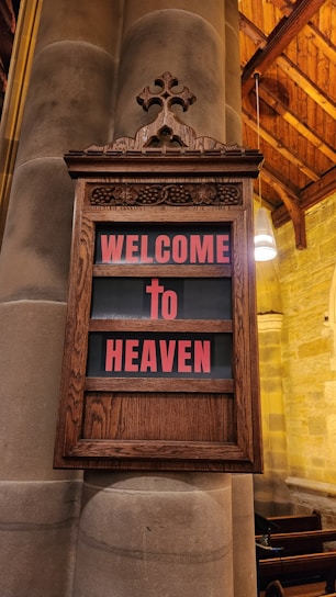 An ornate wooden plaque is mounted on a large stone pillar inside a church. The plaque contains the words 'WELCOME To HEAVEN' in bold red letters. The background is a soft black, and the edges of the plaque feature intricate carvings. The setting suggests a warm, inviting atmosphere with softly lit surroundings and wooden elements.