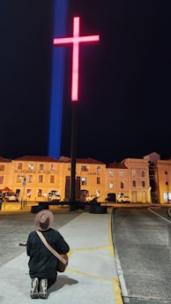 A person kneels on a pathway while holding a guitar, facing a large illuminated red cross. The scene is set at night, with warm light illuminating historic building facades in the background. The atmosphere is calm and contemplative.