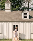 A young woman in a floral dress walks barefoot past a small, white wooden building with a cupola and dormer window. The structure is set against a backdrop of lush green trees and has a rustic appearance. The sunlight casts soft shadows, creating a peaceful, serene atmosphere.