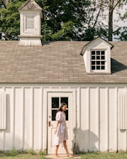 A young woman in a floral dress walks barefoot past a small, white wooden building with a cupola and dormer window. The structure is set against a backdrop of lush green trees and has a rustic appearance. The sunlight casts soft shadows, creating a peaceful, serene atmosphere.