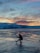 A person is balancing in a crouched position on a frozen, cracked icy surface with a mountainous backdrop. The sky is painted with vibrant hues from a rising or setting sun, adding warmth to the otherwise cold landscape.